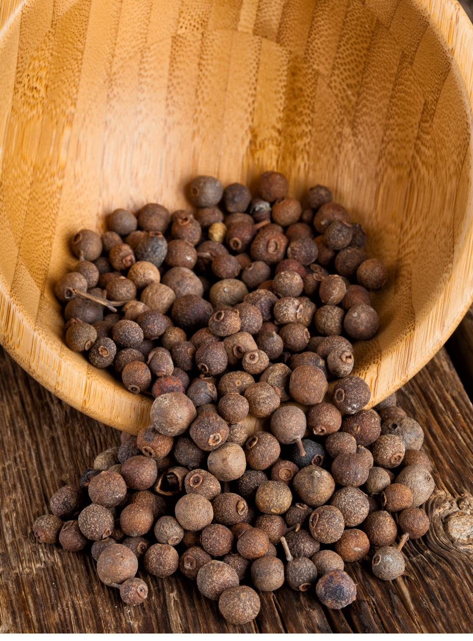 Whole allspice berries spilling from a tipped wooden bowl onto a dark wooden table