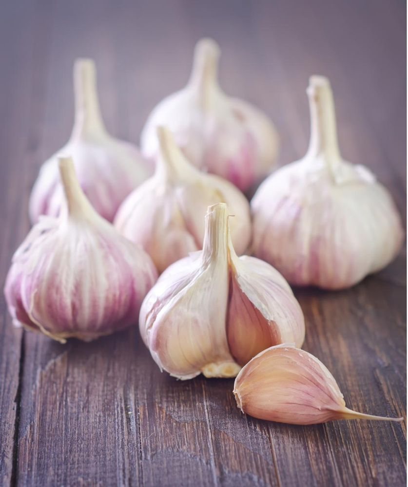 Heads of pink-striped garlic on a dark wooden table, one broken open with a loose clove in front