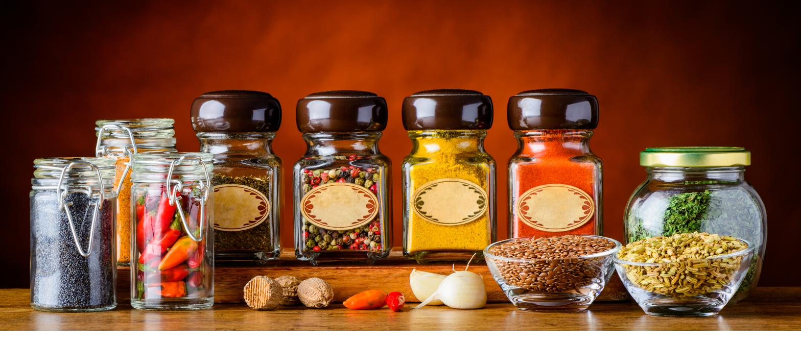 A row of glass spice jars, including mixed peppercorns, turmeric, and paprika, on a wooden shelf against a warm orange wall