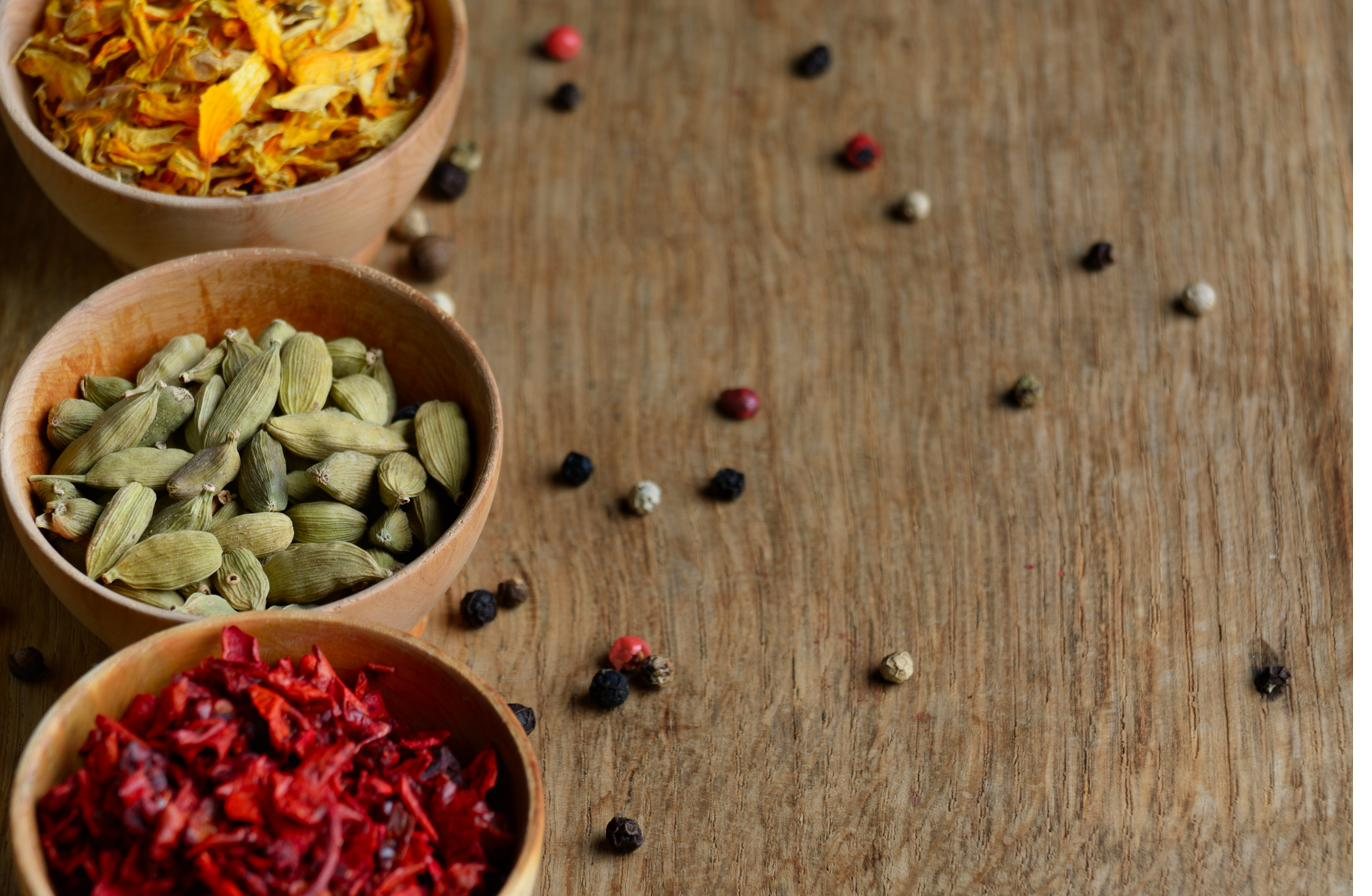 Small bowls of saffron threads, green cardamom pods, and dried flower petals on a wooden table, with peppercorns scattered around